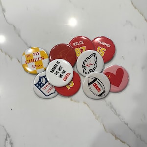 May include: A collection of nine round buttons with various designs related to the Kansas City Chiefs football team. The buttons feature the team's logo, player names and numbers, and phrases like "In My Chiefs Era" and "Karma is the guy on the Chiefs".