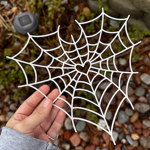 Puede incluir: Corte de papel blanco de una telaraña con un corazón en el centro. La telaraña es intrincada y detallada, con un diseño delicado.