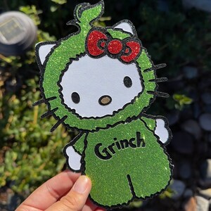 May include: Green glitter Hello Kitty character wearing a Grinch costume with a red bow. The costume has the word "Grinch" written on the front.