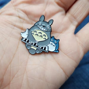Puede incluir: Un pin de esmalte gris y azul con Totoro, un personaje de la pel&iacute;cula animada Mi vecino Totoro, tocando el piano. El pin es un objeto de colecci&oacute;n popular para los fans de la pel&iacute;cula.