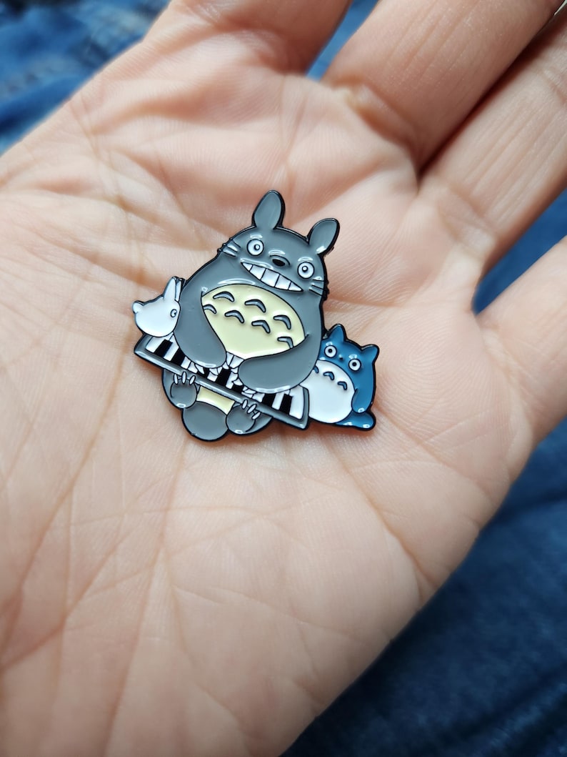 Puede incluir: Un pin de esmalte gris y azul con Totoro, un personaje de la pel&iacute;cula animada Mi vecino Totoro, tocando el piano. El pin es un objeto de colecci&oacute;n popular para los fans de la pel&iacute;cula.