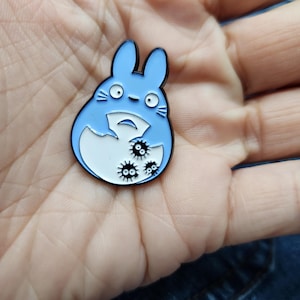 Puede incluir: Un pin de esmalte azul con un personaje de dibujos animados con barriga blanca y ojos negros. El personaje sostiene un huevo blanco con tres criaturas negras y espinosas en su interior.