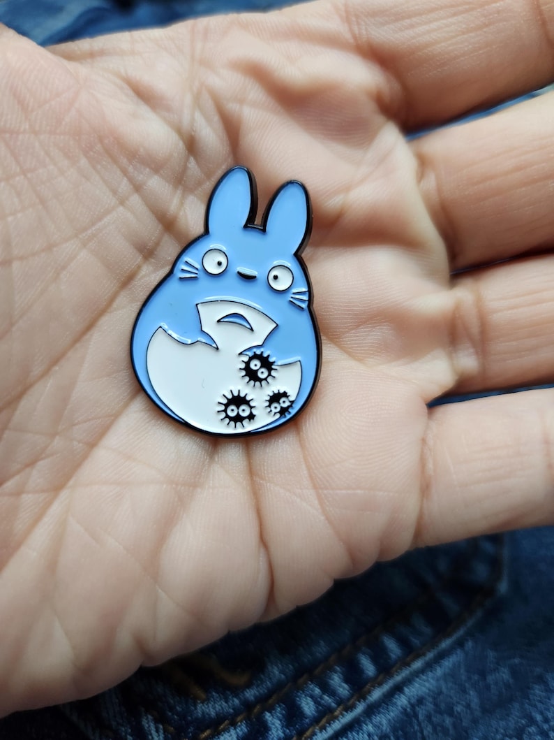 Puede incluir: Un pin de esmalte azul con un personaje de dibujos animados con barriga blanca y ojos negros. El personaje sostiene un huevo blanco con tres criaturas negras y espinosas en su interior.