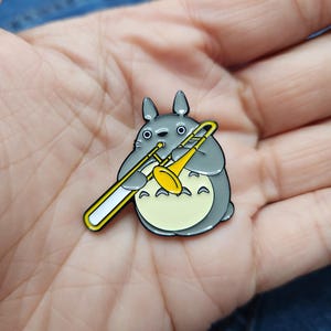Puede incluir: Un pin de esmalte gris y amarillo con un personaje de dibujos animados tocando un tromb&oacute;n. El personaje tiene ojos grandes y redondos y una boca peque&ntilde;a. El tromb&oacute;n es amarillo con un tobog&aacute;n blanco.