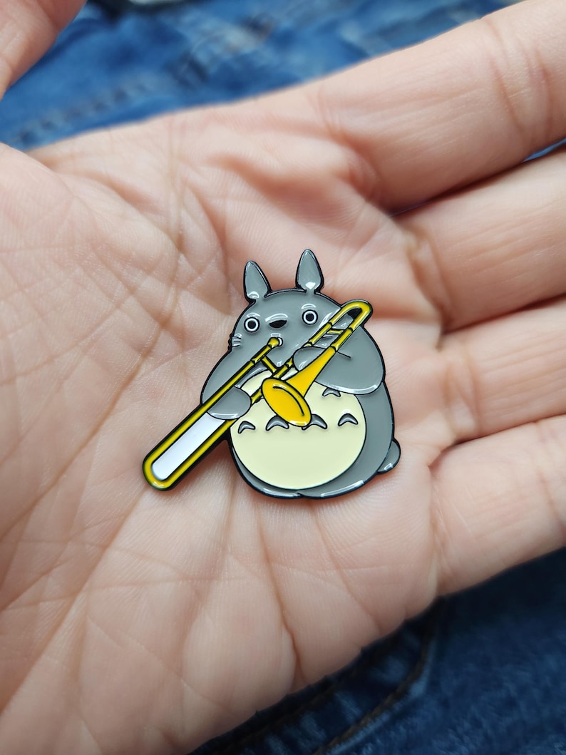 Puede incluir: Un pin de esmalte gris y amarillo con un personaje de dibujos animados tocando un tromb&oacute;n. El personaje tiene ojos grandes y redondos y una boca peque&ntilde;a. El tromb&oacute;n es amarillo con un tobog&aacute;n blanco.