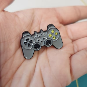 May include: A gray metal video game controller pin with yellow buttons. The pin is a replica of a classic video game controller.