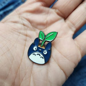 Puede incluir: Un pin de esmalte azul y blanco con un personaje de dibujos animados con una planta verde que le crece de la cabeza.