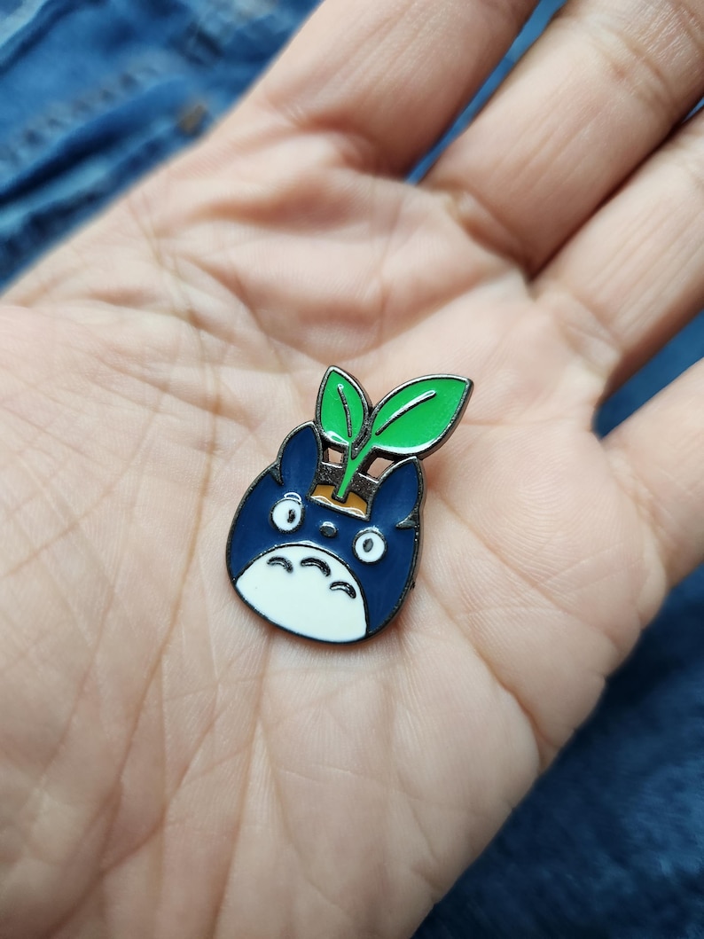 Puede incluir: Un pin de esmalte azul y blanco con un personaje de dibujos animados con una planta verde que le crece de la cabeza.