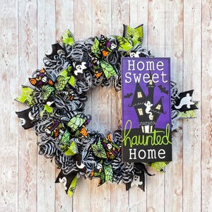 May include: A Halloween wreath featuring black and white mesh, lime green spider web ribbons, and patterned fabric with ghosts and pumpkins. A rectangular purple sign is attached, displaying a haunted house illustration and the text Home Sweet Haunted Home.
