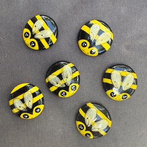 May include: Six circular, smooth, black and yellow striped bee-themed cabochons. Each piece features a painted face with two large black eyes, a small smile, and two glittery, translucent, light-colored wings positioned on the upper back section.