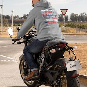 Op de afbeelding: Een persoon op een zwarte motorfiets, gekleed in een grijze hoodie met de tekst "AMERICAN SPEED" en "FIGHT FOR FREEDOM" met een autografiek. De motorfiets heeft een kentekenplaat met "CH8". De rijder draagt een blauwe spijkerbroek en bruine laarzen.