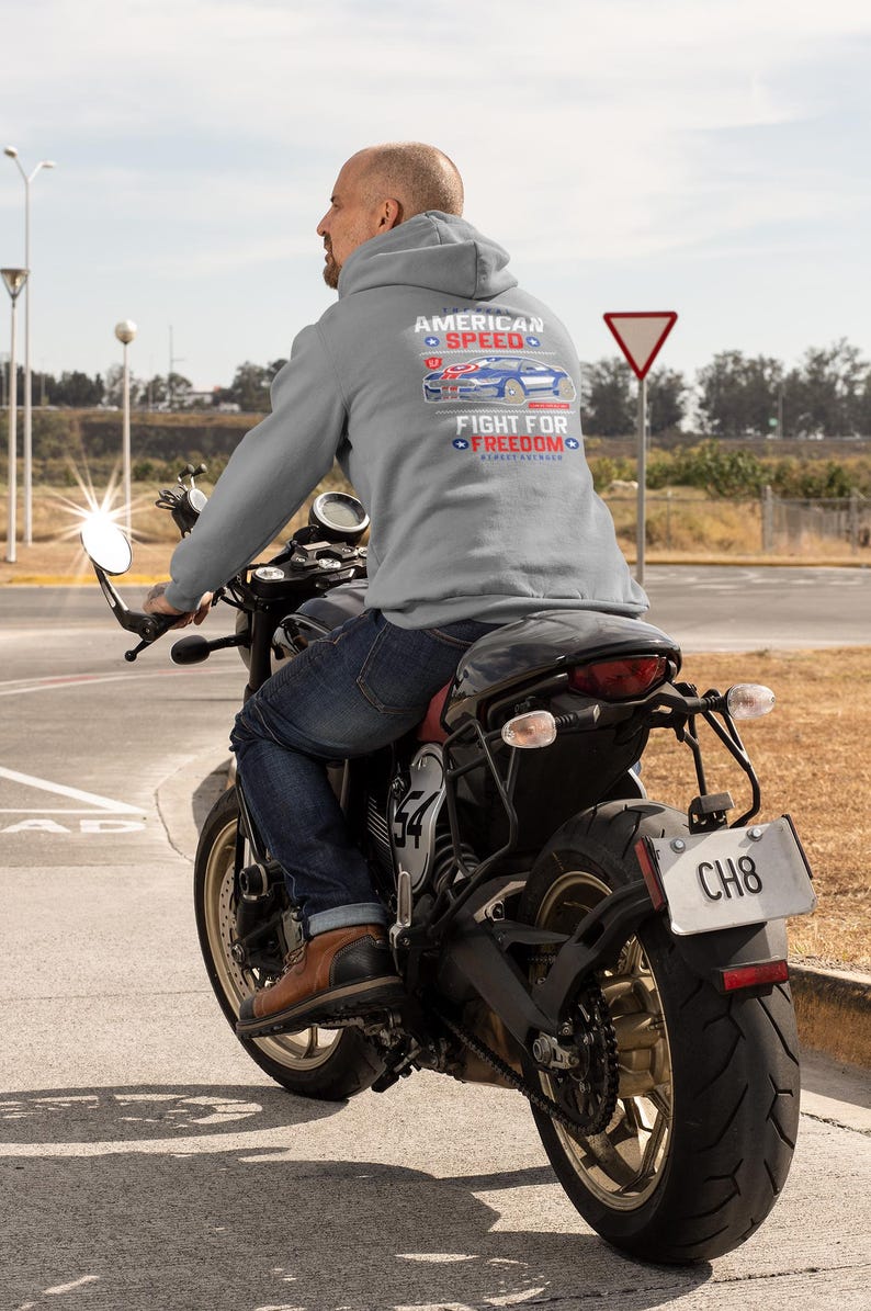 Op de afbeelding: Een persoon op een zwarte motorfiets, gekleed in een grijze hoodie met de tekst "AMERICAN SPEED" en "FIGHT FOR FREEDOM" met een autografiek. De motorfiets heeft een kentekenplaat met "CH8". De rijder draagt een blauwe spijkerbroek en bruine laarzen.