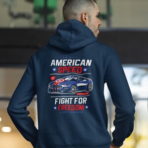 Op de afbeelding: Marineblauwe hoodie met een afbeelding van een blauwe sportwagen en de tekst "AMERICAN SPEED" en "FIGHT FOR FREEDOM". Het ontwerp bevat rood, wit en blauw, wat een patriottisch thema oproept. De hoodie is een casual kledingstuk.