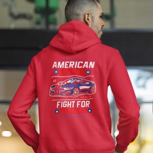 Op de afbeelding: Rode hoodie met een afbeelding op de rug met een blauwe en rode sportwagen, de tekst "AMERICAN SPEED" en "FIGHT FOR FREEDOM". De hoodie heeft een capuchon en lange mouwen.