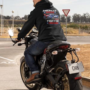 Op de afbeelding: Een zwarte hoodie met de tekst "AMERICAN SPEED" en "FIGHT FOR FREEDOM" met een rode en blauwe autografiek. De persoon rijdt op een zwarte motorfiets op een verharde weg.