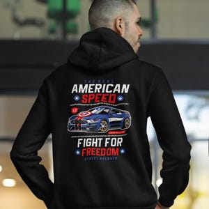 Op de afbeelding: Zwarte hoodie met een afbeelding van een blauwe sportwagen en de tekst "The Real American Speed" en "Fight for Freedom". Het ontwerp bevat rode, witte en blauwe elementen, die een patriottisch thema oproepen.