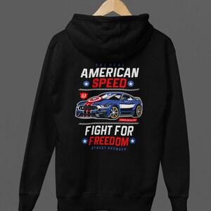 Op de afbeelding: Zwarte hoodie met een afbeelding van een blauwe sportwagen, rode en witte strepen en tekst. Het ontwerp bevat de zinnen "THE REAL AMERICAN SPEED" en "FIGHT FOR FREEDOM" met sterren, in een patriottische stijl.