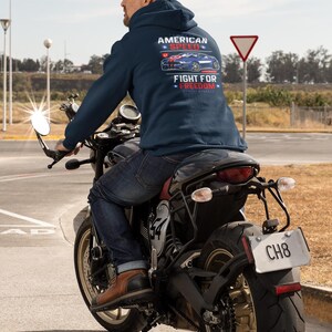 Op de afbeelding: Een persoon op een motor draagt een marineblauwe hoodie met de tekst "AMERICAN SPEED" en "FIGHT FOR FREEDOM" boven een autografiek. De persoon draagt een blauwe spijkerbroek en bruine laarzen.
