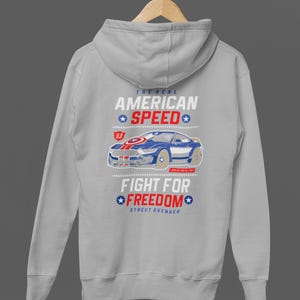 Op de afbeelding: Een lichtgrijze hoodie met een afbeelding van een blauwe sportwagen en de tekst "THE REAL AMERICAN SPEED" en "FIGHT FOR FREEDOM". Het ontwerp bevat rode en blauwe elementen. De hoodie heeft een capuchon en lange mouwen.