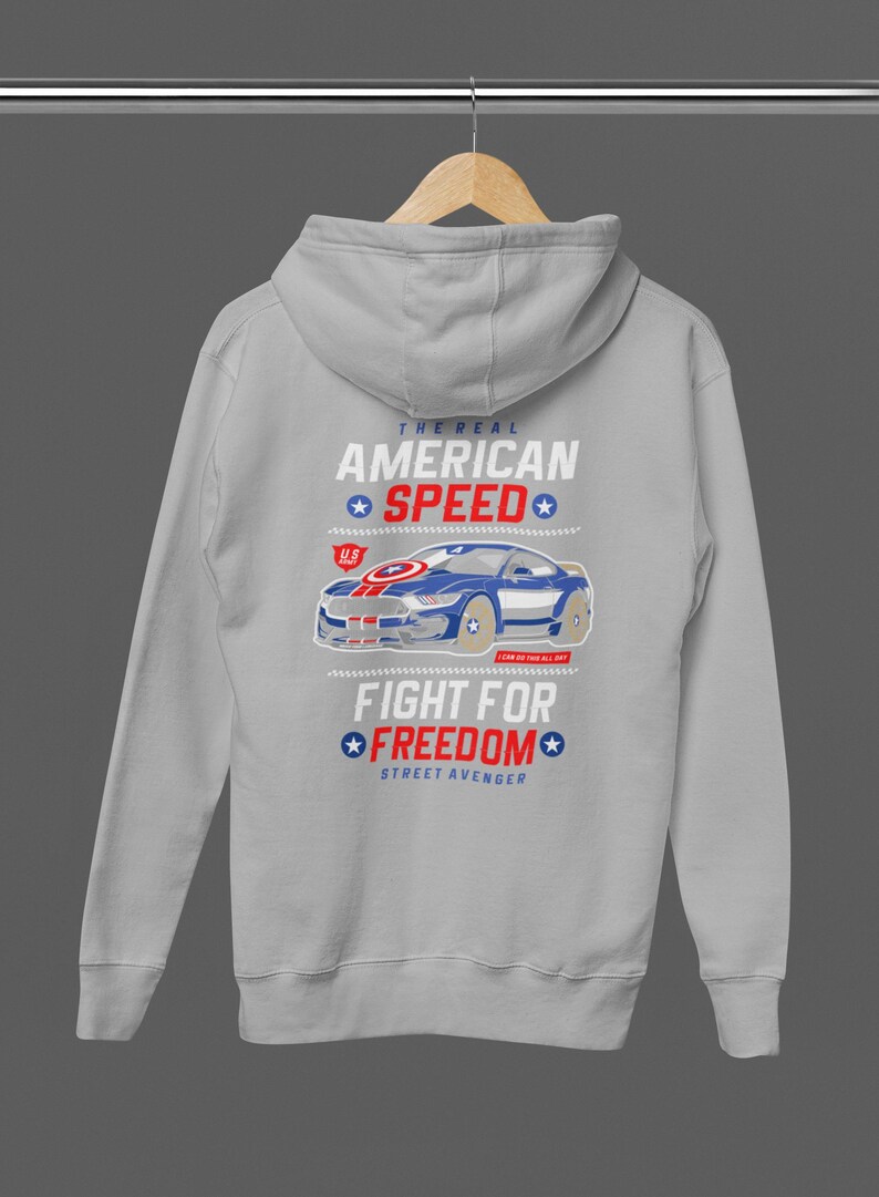 Op de afbeelding: Een lichtgrijze hoodie met een afbeelding van een blauwe sportwagen en de tekst "THE REAL AMERICAN SPEED" en "FIGHT FOR FREEDOM". Het ontwerp bevat rode en blauwe elementen. De hoodie heeft een capuchon en lange mouwen.