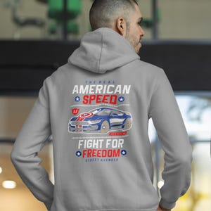 Op de afbeelding: Een grijze hoodie met een afbeelding op de achterkant. De afbeelding toont een blauwe en witte auto met de tekst "The Real American Speed" en "Fight for Freedom". De hoodie is een casual stijl.