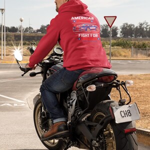 Op de afbeelding: Een persoon op een zwarte motorfiets draagt een rode hoodie met de tekst "AMERICAN FIGHT FOR" en een autografiek. De motorfiets heeft een kentekenplaat met "CH8". De persoon draagt een blauwe spijkerbroek en bruine laarzen.