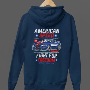 Op de afbeelding: Marineblauwe hoodie met een afbeelding van een blauwe sportwagen en de tekst "AMERICAN SPEED" en "FIGHT FOR FREEDOM". Het ontwerp bevat rood-witte strepen op de auto en steraccenten.
