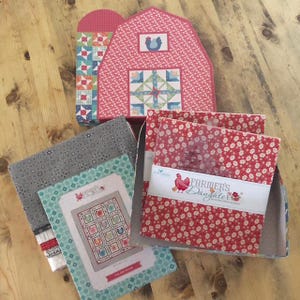 May include: A collection of quilting supplies, including fabric squares and a barn-shaped box. The box is pink with a rooster design and a patchwork pattern. The fabric squares are red with white floral patterns. A label reads "Farmer's Daughter".