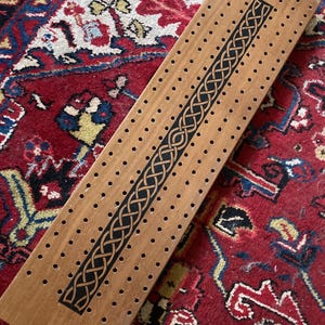 May include: A rectangular wooden cribbage board featuring a central black panel decorated with an intricate, repeating Celtic knot pattern. The board has three parallel rows of small, evenly spaced holes drilled into the light brown wood surface.