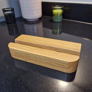 May include: A light oak wooden phone stand with a slot for a phone. The stand has a smooth, curved design. The stand is approximately 15 cm long and 7.6 cm wide. The wood grain is visible.