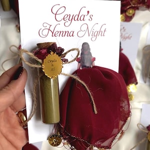 Może przedstawiać: Mała szklana butelka wypełniona zielonym proszkiem henny, związana sznurkiem i małym bukietem sztucznych kwiatów. Butelka jest przymocowana do burgundowego jedwabnego szala z złotą lamówką i monetami. Karta zawiera napis "Ceyda's Henna Night" z imionami "Ceyda & Ali" na złotej pieczęci.