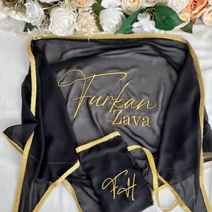 May include: Black sheer shawl and matching pouch with gold trim and embroidery. The shawl features the name "Furkan Zava" in gold script, and the pouch has the initials "FH". The items are displayed on a white surface with floral accents.