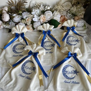 May include: Cream-colored fabric bags with blue and gold ribbons, each embroidered with a crescent moon, star, and the name "Kadir". The bags are decorated with a blue eye charm and are arranged on a white surface.