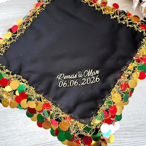 May include: Black square textile with gold trim and colorful sequin fringe. The textile has embroidered text in gold thread. The trim is gold with red, green, and gold sequins.