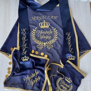May include: Navy blue ceremonial outfit with gold embroidery. The set includes a cape, sash, and small bag, all adorned with gold text and crown designs. The cape and bag feature the name "Mustafa Yagiz" in elegant script. The sash has the name "Mustafa" and "Yagiz" on each side.