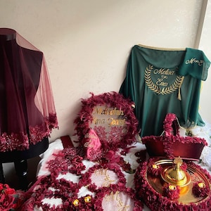 May include: A burgundy and gold wedding ceremony set with a veil, a decorative pillow, a tray with a golden teapot, and two floral wreaths with the letters "M & F" embroidered on them.