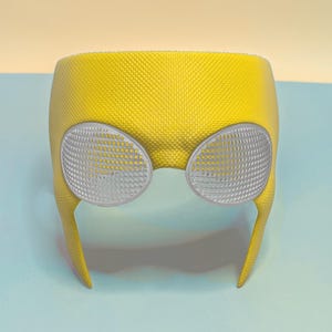 May include: A bright yellow mask featuring a textured, honeycomb-like surface pattern and two large, oval-shaped silver eye lenses. The lenses have a fine, perforated grid design, and the mask has a curved, rigid structure that covers the upper face.