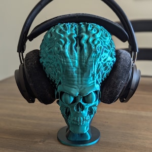 May include: A teal blue 3D printed skull headphone stand with a detailed brain design. The stand is holding a pair of black over-ear headphones.
