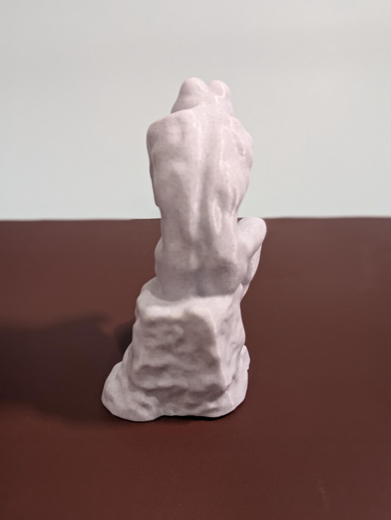 May include: A white ceramic sculpture of a frog sitting on a rock. The frog is in a seated position with its legs crossed and its head tilted down. The sculpture is detailed with textured surfaces and a smooth finish.