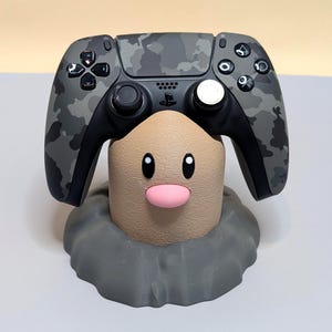 May include: A gray camouflage video game controller rests on a tan, cylindrical character stand with a pink nose and black eyes. The character is set within a textured, dark gray base that resembles a mound of earth or rock, providing a stable mount.