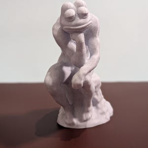 May include: A white 3D printed sculpture of a frog in a thinking pose, similar to the famous "The Thinker" statue. The frog is sitting on a small base.