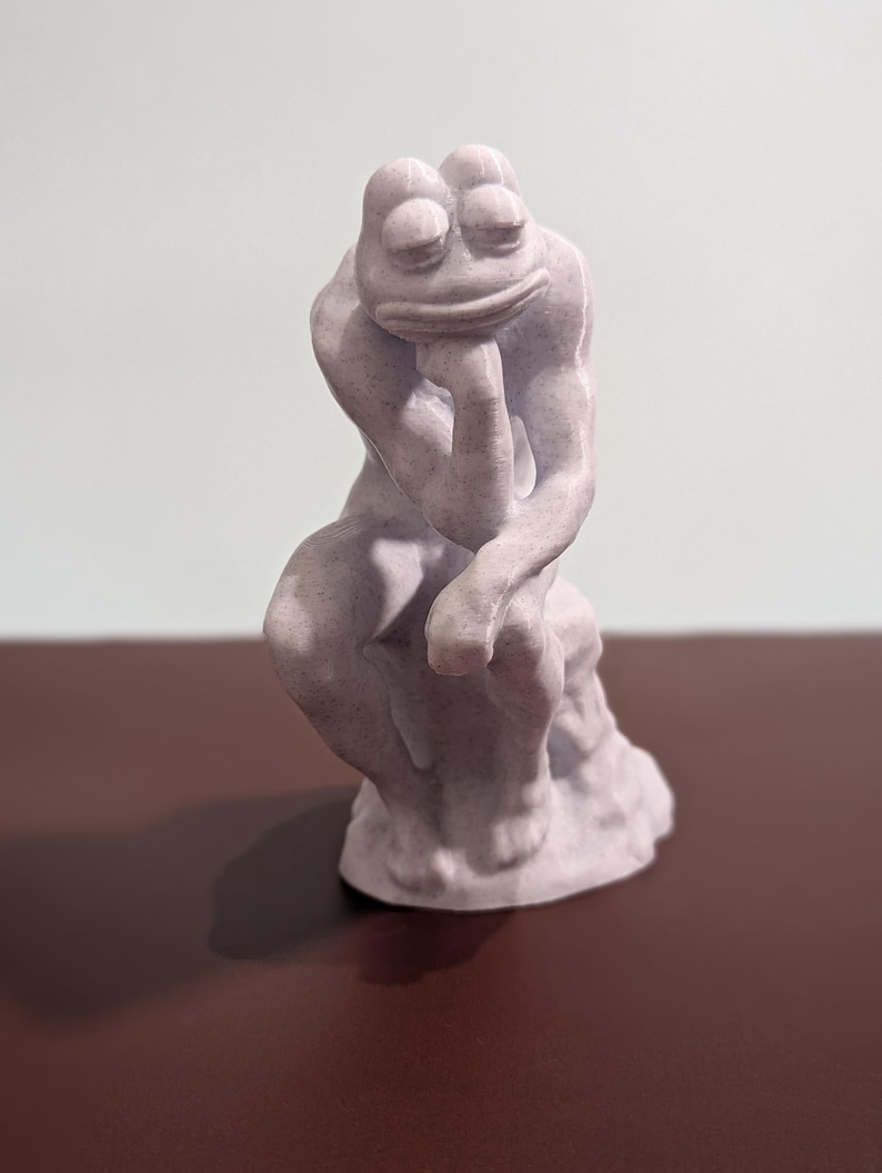 May include: A white 3D printed sculpture of a frog in a thinking pose, similar to the famous "The Thinker" statue. The frog is sitting on a small base.