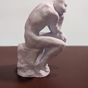 May include: A small, gray, 3D printed sculpture of a person sitting with their chin resting on their hand, in a contemplative pose. The sculpture is on a textured base.