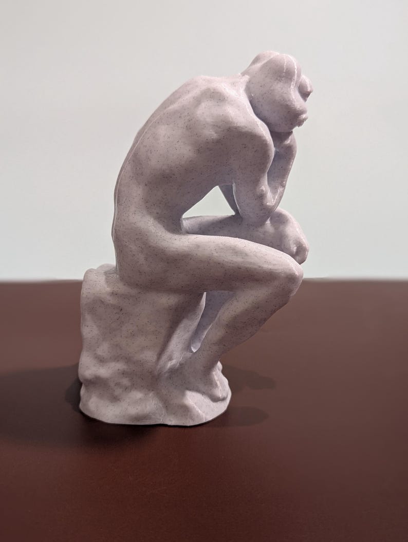 May include: A small, gray, 3D printed sculpture of a person sitting with their chin resting on their hand, in a contemplative pose. The sculpture is on a textured base.
