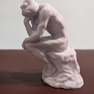 May include: A 3D printed sculpture of a person sitting in a contemplative pose, resembling the famous "Thinker" statue. The sculpture is made of a light gray, textured material and has a slightly distressed finish.