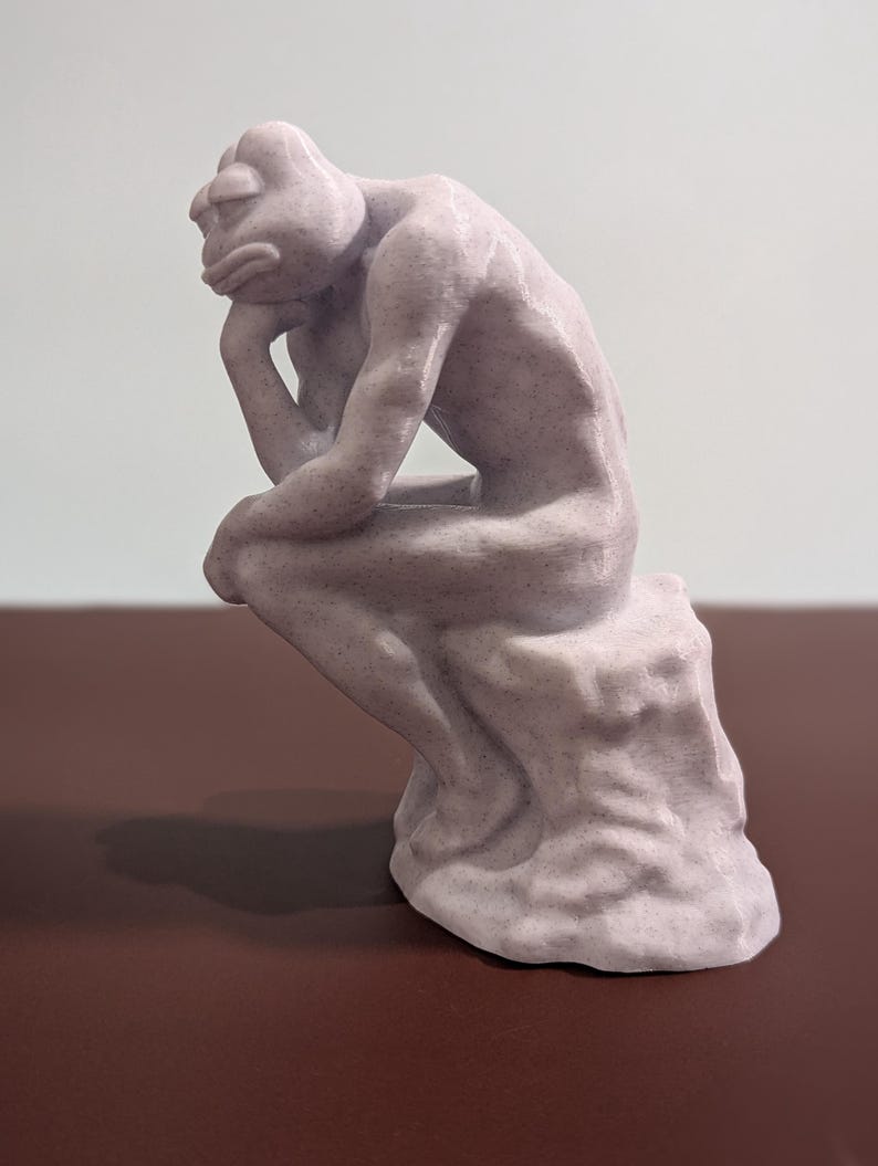 May include: A 3D printed sculpture of a person sitting in a contemplative pose, resembling the famous "Thinker" statue. The sculpture is made of a light gray, textured material and has a slightly distressed finish.