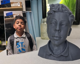 May include: A dark gray 3D printed bust of a young person with short hair, positioned next to a rectangular photograph of the same individual wearing a gray t-shirt with the text ST. AN printed in blue letters and a black backpack.