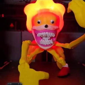 May include: A yellow, illuminated creature figure with a large head, glowing orange top, and a wide pink mouth filled with numerous sharp, white teeth. The figure has two small black eyes, thin yellow limbs, and one visible red shoe on a foot.