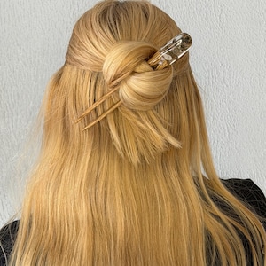 May include: A blonde hairstyle with a bun secured by a wooden hair stick. The hair stick has a clear resin top with embedded floral elements. The hair is long and flowing, with a natural, sun-kissed color.