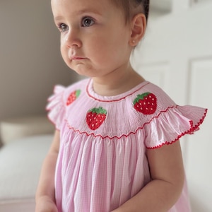 May include: A little girl wearing a pink and white gingham dress with embroidered strawberries. The dress has red trim and short sleeves.